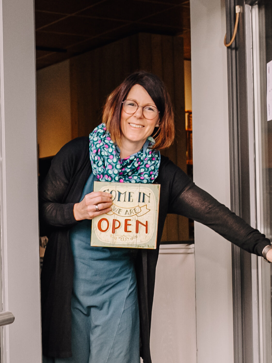 Die Inhaberin Martina schaut freudig und lächelnd in die Kamera, guckt dabei aus der Eingangstür heraus und hält ein Schild auf dem "Come In, We Are Open" steht.
