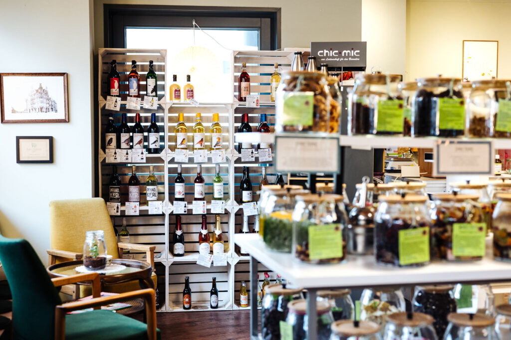 Im Hintergrund sind Weinkisten zu einem Regal gestapelt sind un in dem sich unterschiedliche Weiß- und Rotweine befinden. Im Vordergrund sind unscharf Behälter mit Süßigkeiten. Links sind gepolsterte Sessel 