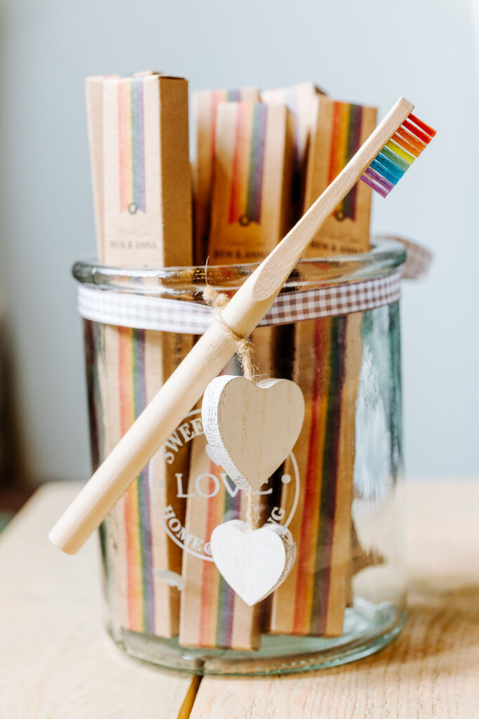 Eine hölzerne Regenbogenzahnbürste die einem Glas hängt. Dahinter sind Packungen der Regenbogen Zahnbürsten