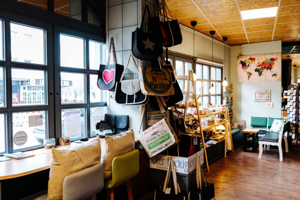 Blick auf gepolsterte Barhocker mit Kissen an einer Theke. Im Hintergrund ist eine Eckbank und ein Beistelltisch aus zwei Getränkekisten. Zwischen den Barhockern und der Sitzecke ist eine Säule, an der unterschiedliche EInkaufstaschen hängen.