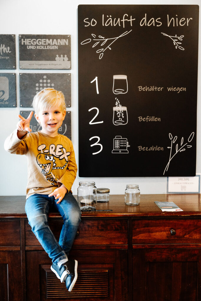 Ein Kind mit gelben Pullover, auf welchem ein T-Rex abgebildet ist, sitzt auf einer Theke aus Holz und zeigt drei Finger. Im Hintergrund ist eine Tafel abgebildet mit drei Schritten. Darüber steht "so läuft das hier"
1. Behählter wiegen
2. Befüllen
3. Bezhalen