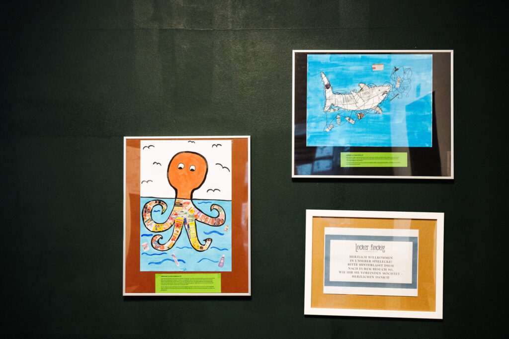 Wand mit drei Fotos. Einem Oktopus welch zur Hälfte aus dem Meer schaut und die Tentakel mit Plastikmüll ausgefüllt sind. Daneben ist ein Motiv eines Hais, welch sich in einem Netz mit Müll verfangen hat.