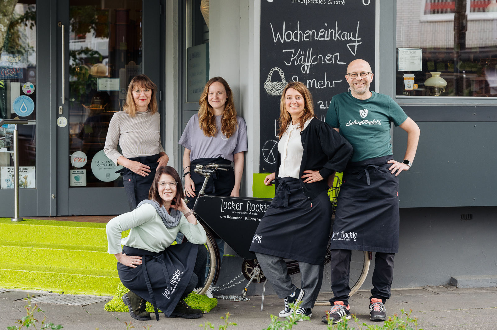 Gruppenbild von mehreren Personen vor einem Fahrrad. Alle tragen Schürzen auf denen Locker Flockig steht. Die Gruppe befindet sich vor dem Eingang des Geschäftes.