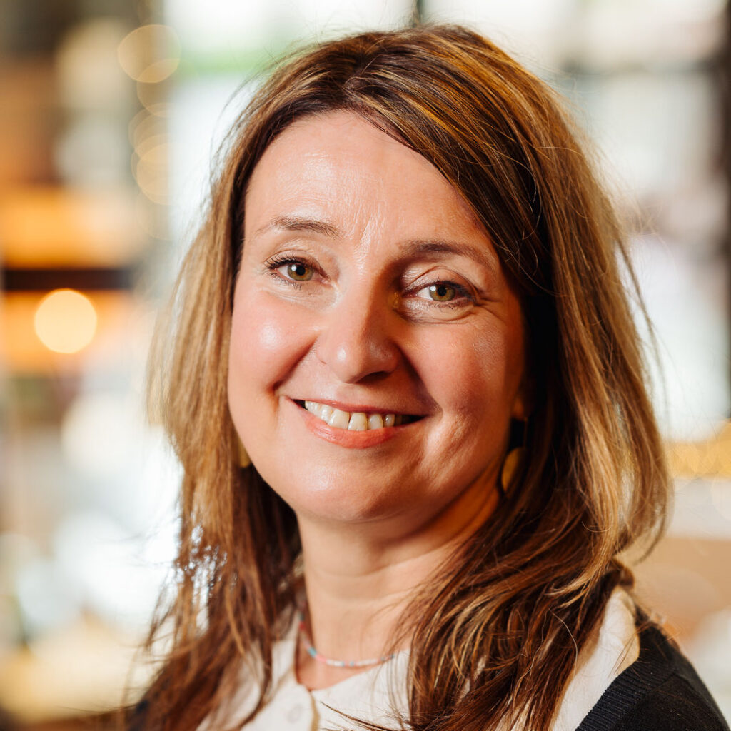 Potraitfoto einer braunhaarigen Person mit längeren haaren, die lächelt in die Kamera schaut.
