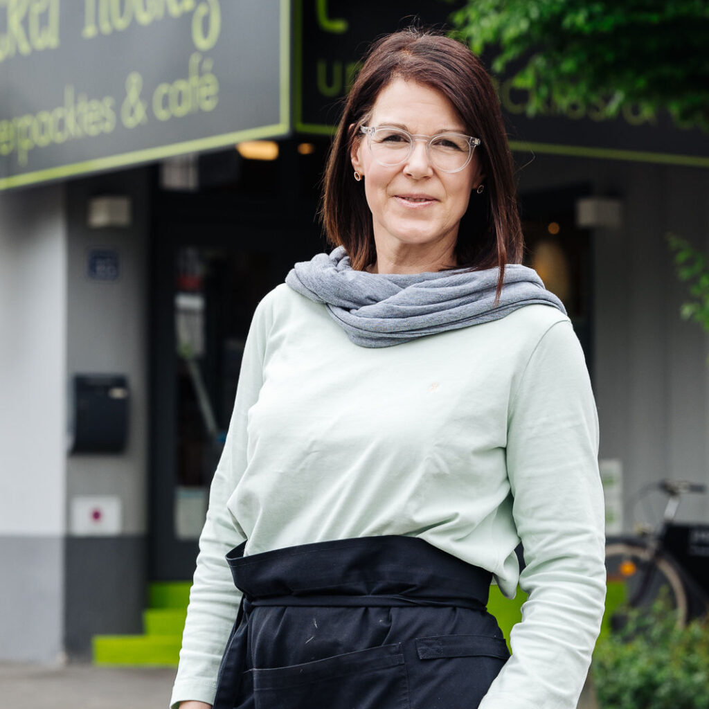 Potraitfoto der Inhaberin Martina mit dunkelbraunen langen Haar, welch lächelnd in die Kamera schaut. Martina steht vor dem Geschäft und trägt einen grauen Schal und eine Schürze