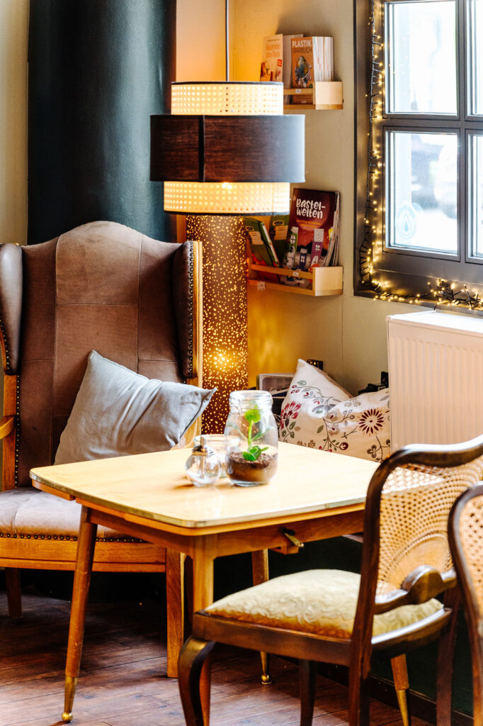 Foto einer gemütlichen Sitzecke in einem Café mit Holzstuhl, Ledersessel und einem Holztisch auf dem ein Glas mit Pflanze in Erde sich befindet. Das Fenster ist mit einer Lichterkette dekoriert.