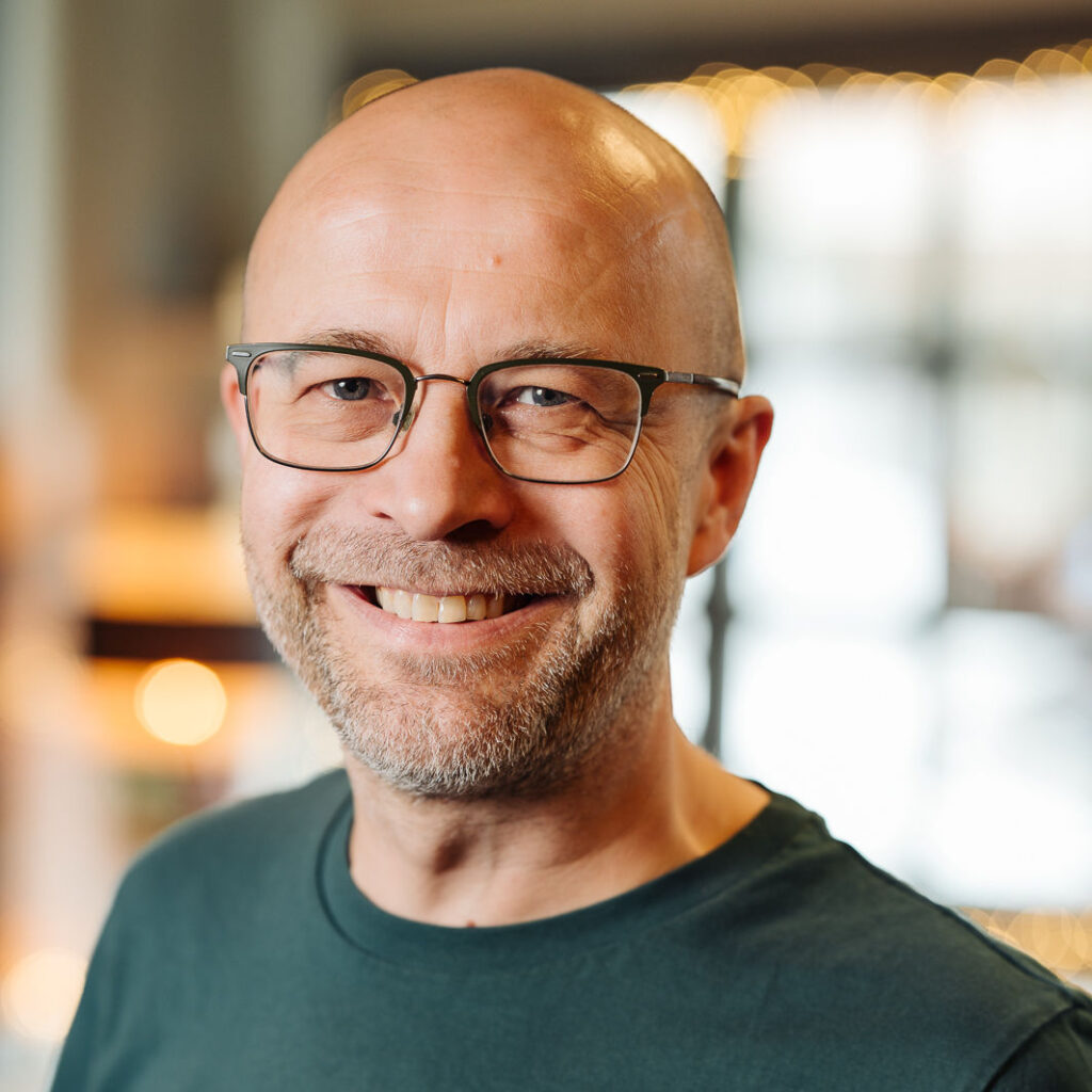 Potraitfoto einer Person mit dreitage Bart, Brille und Glatze, welch lächelnd in die Kamera schaut.