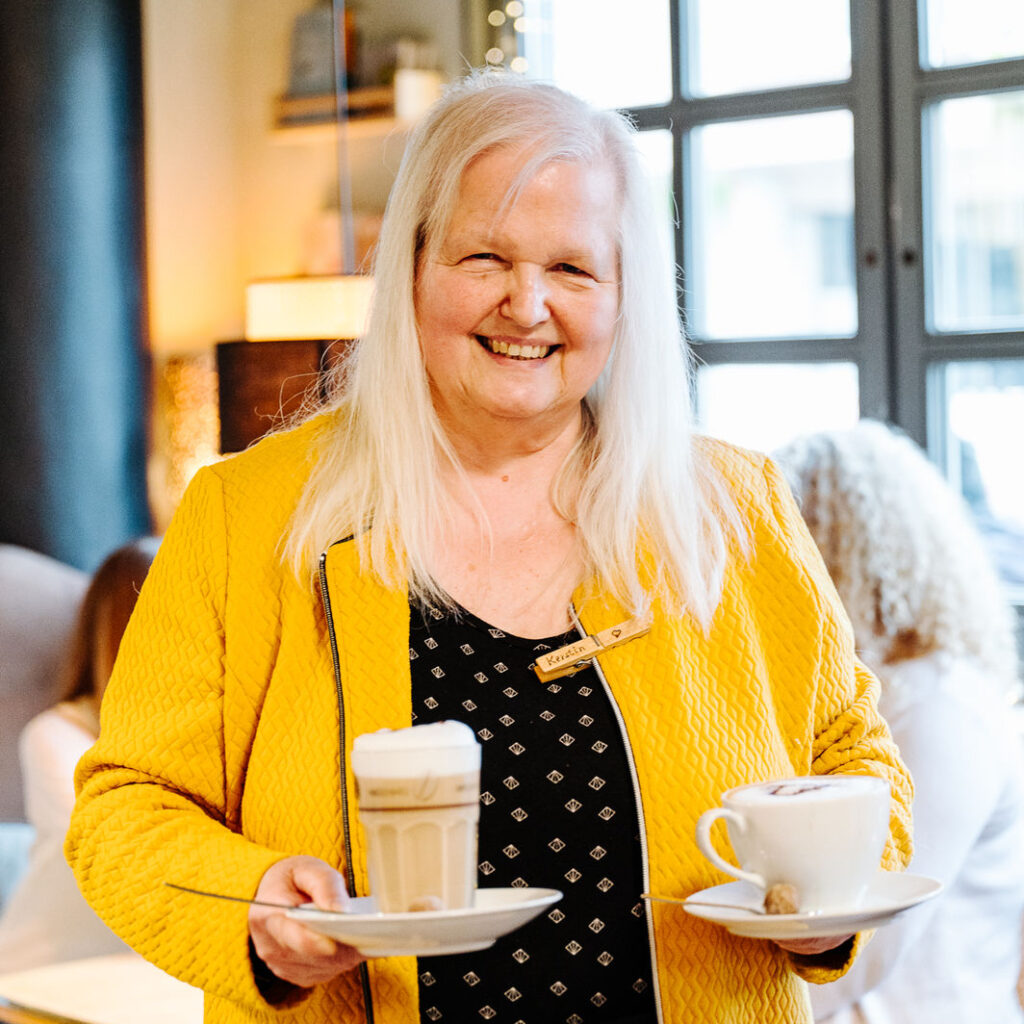 Potraitfoto einer Person mit längeren grauen Haaren und gelber Jacke, die freudig in die Kamera schaut und zwei Heißgetränke hält.