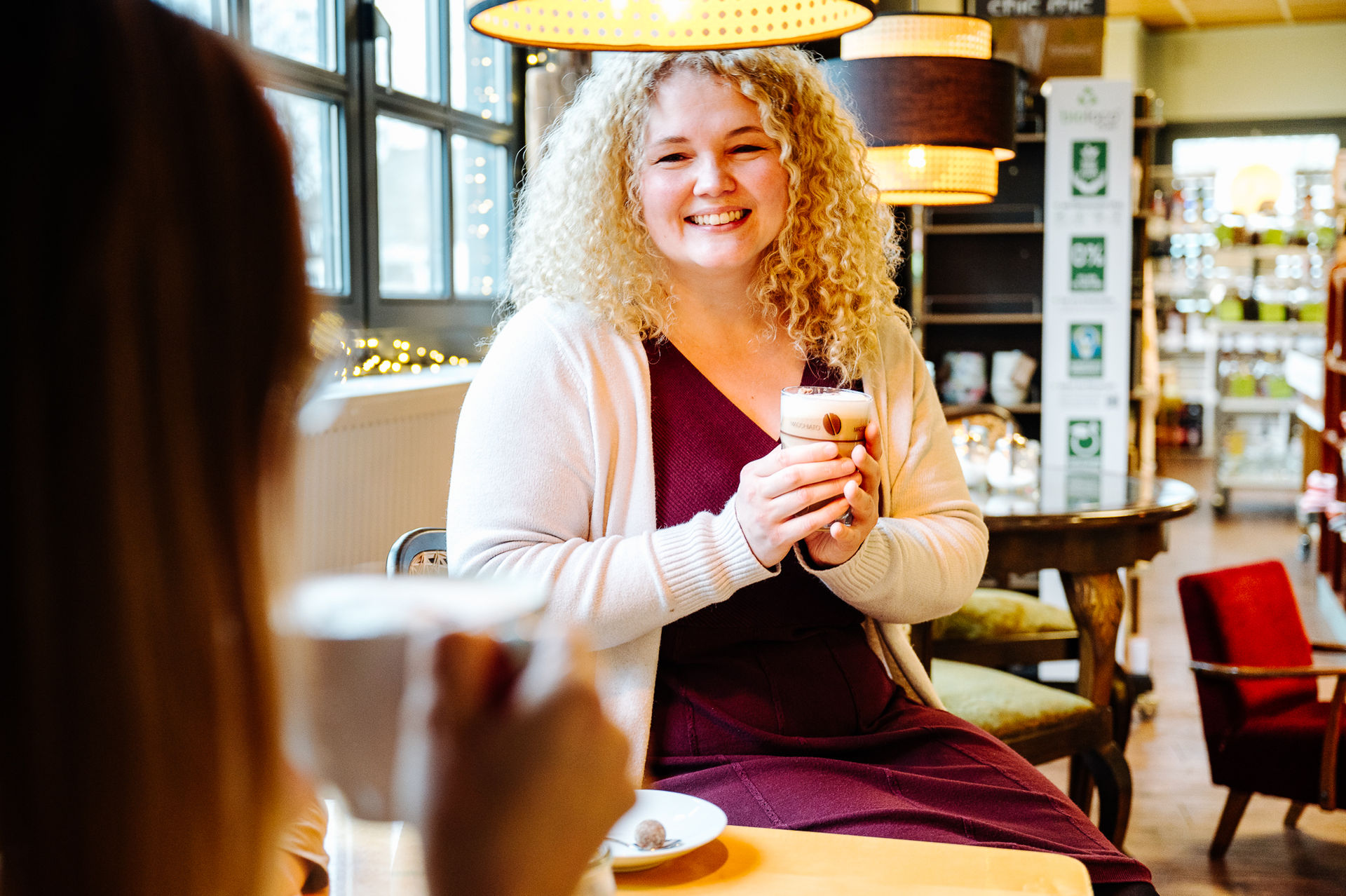 Eine Person sitzt im Café, hat lockiges blondes langes Haar und lächelt eine Person an, welche von Hinten Unscharf im Vordergrund befindet. Beide Personen halten ein Heißgetränk in der Hand